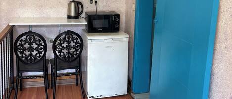 Una kitchenette compacta con microondas, hervidor de agua y un refrigerador pequeño. Hay dos sillas con respaldo decorado. Se ve una puerta azul a la derecha.