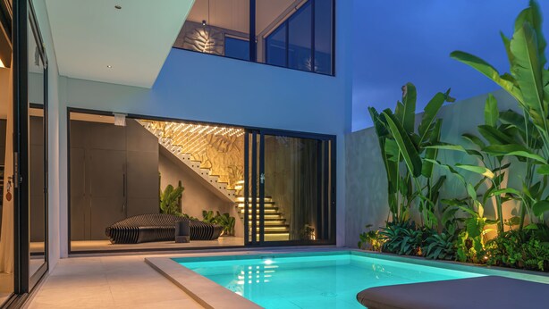 Un área de piscina moderna al aire libre con una escalera que conduce a un segundo piso, rodeada de exuberante vegetación.