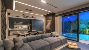 Una sala de estar moderna con un sofá seccional gris, un comedor y una cocina. Hay un ventanal con vista a una piscina.