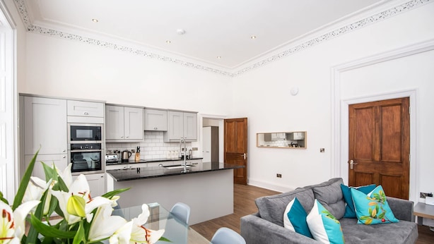 A modern kitchen with white cabinets, a central island, and a dining area with a glass table and chairs. - The Montague (Durham)
