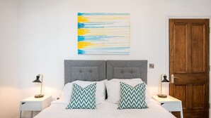 A neatly made bed with two pillows, a wall-mounted abstract painting, and a wooden door. - The Montague (Durham)