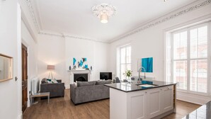A spacious living area with a fireplace, grey sofas, a coffee table, and a kitchen island with a sink. - The Montague (Durham)