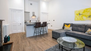 A modern kitchen with a central island, two bar stools, and a grey sofa with patterned cushions. - The Morton (Durham)