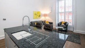 A modern kitchen with a granite countertop, a built-in sink, and a dish rack. There is a grey sofa with yellow cushions, a small round table, and a lamp. A large window with blinds is visible. - The Morton (Durham)