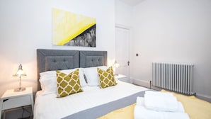 A neatly made bed with patterned pillows, a bedside lamp, and a radiator. - The Morton (Durham)