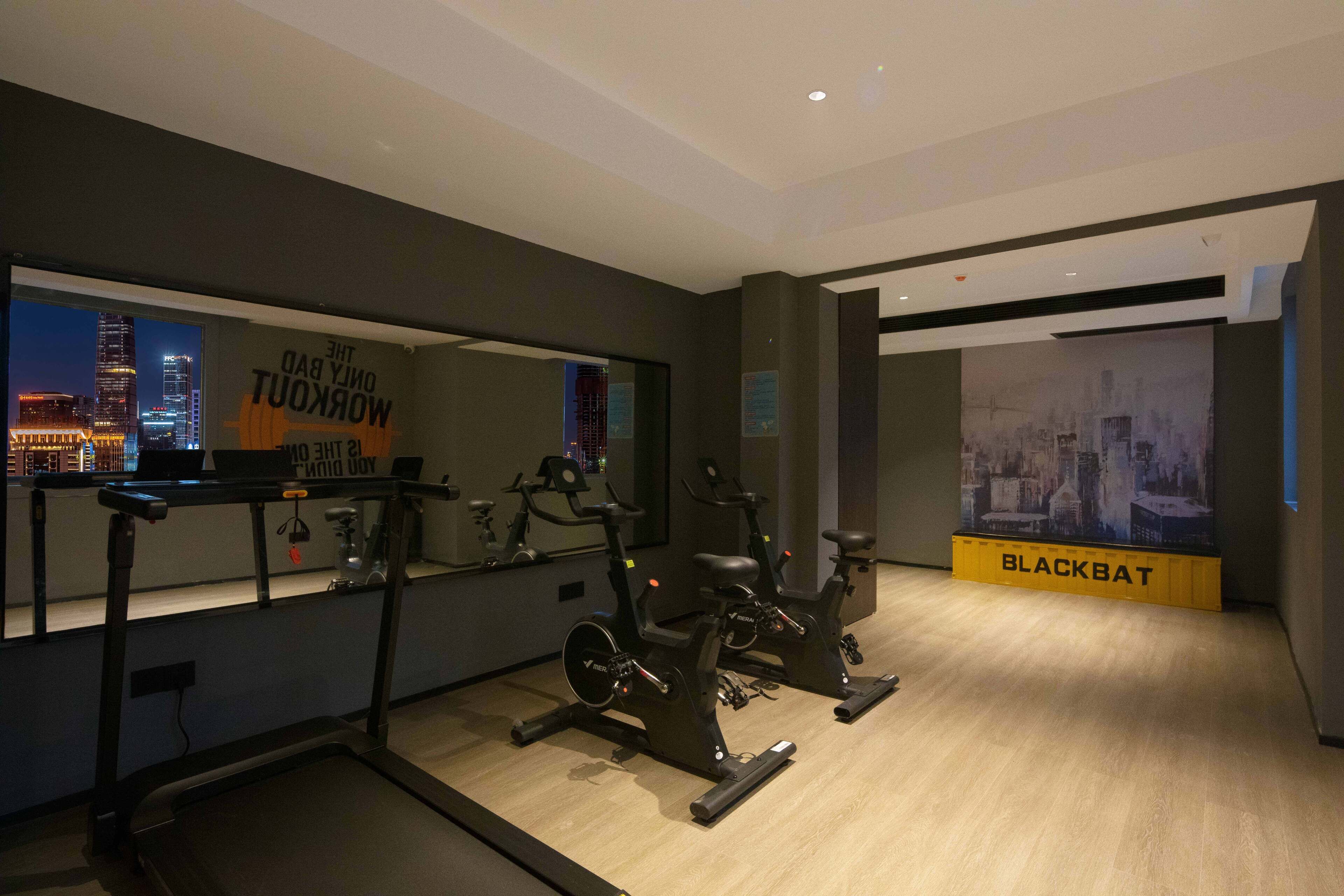 A gym with exercise equipment, a treadmill, and a mirrored wall reflecting a cityscape.