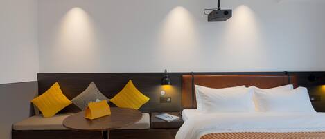 A modern hotel room with a bed, a sofa, a small table, and a wall-mounted projector.