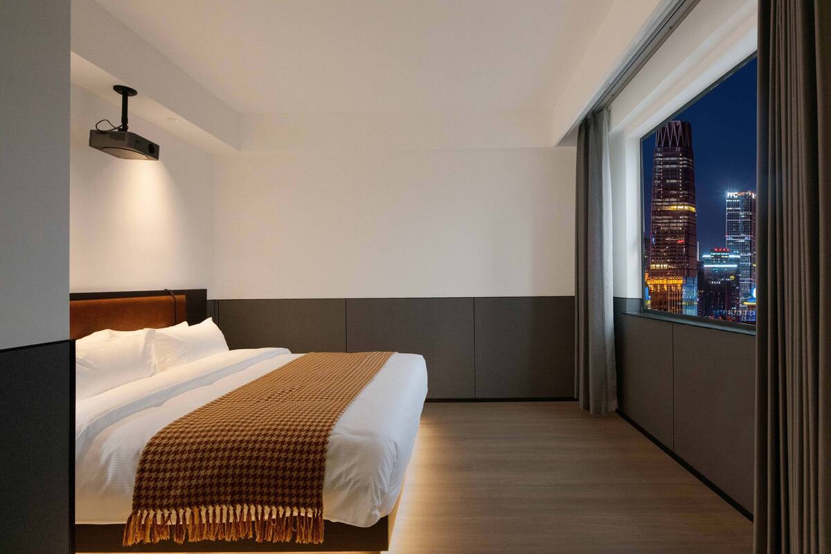 Ein modernes Hotelzimmer mit einem großen Bett, Blick auf die Stadt und einem Projektor an der Decke.