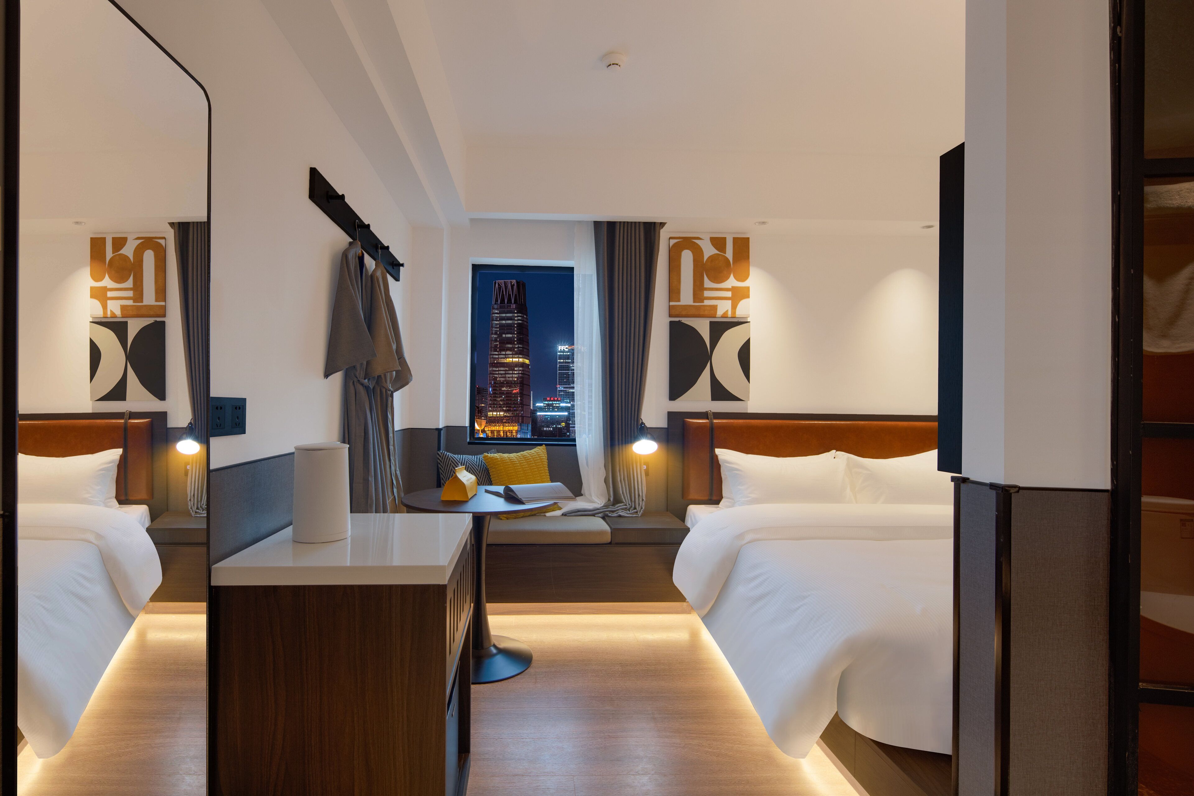 Habitación de hotel con dos camas, un escritorio, una silla, un espejo y vistas a la ciudad iluminada de noche.