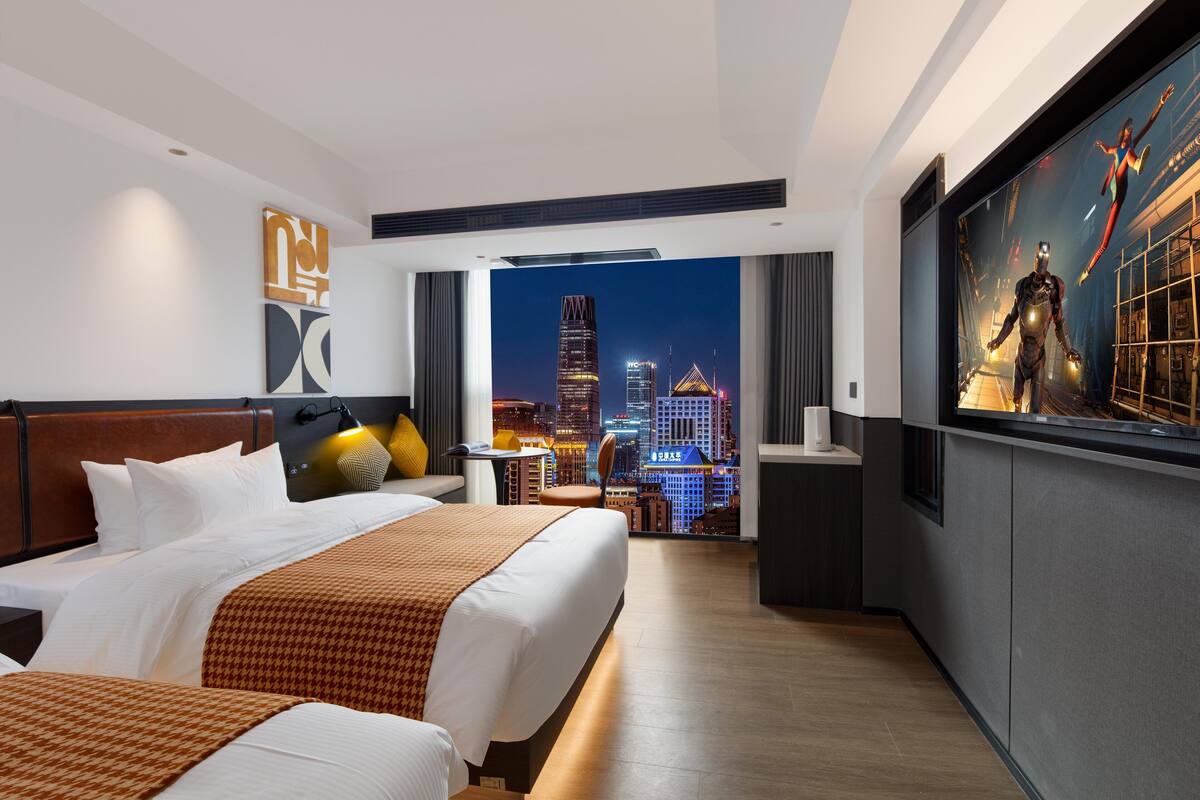 Ein Hotelzimmer mit einem großen Bett, einem Schreibtisch und Blick auf eine nächtliche Stadtlandschaft.