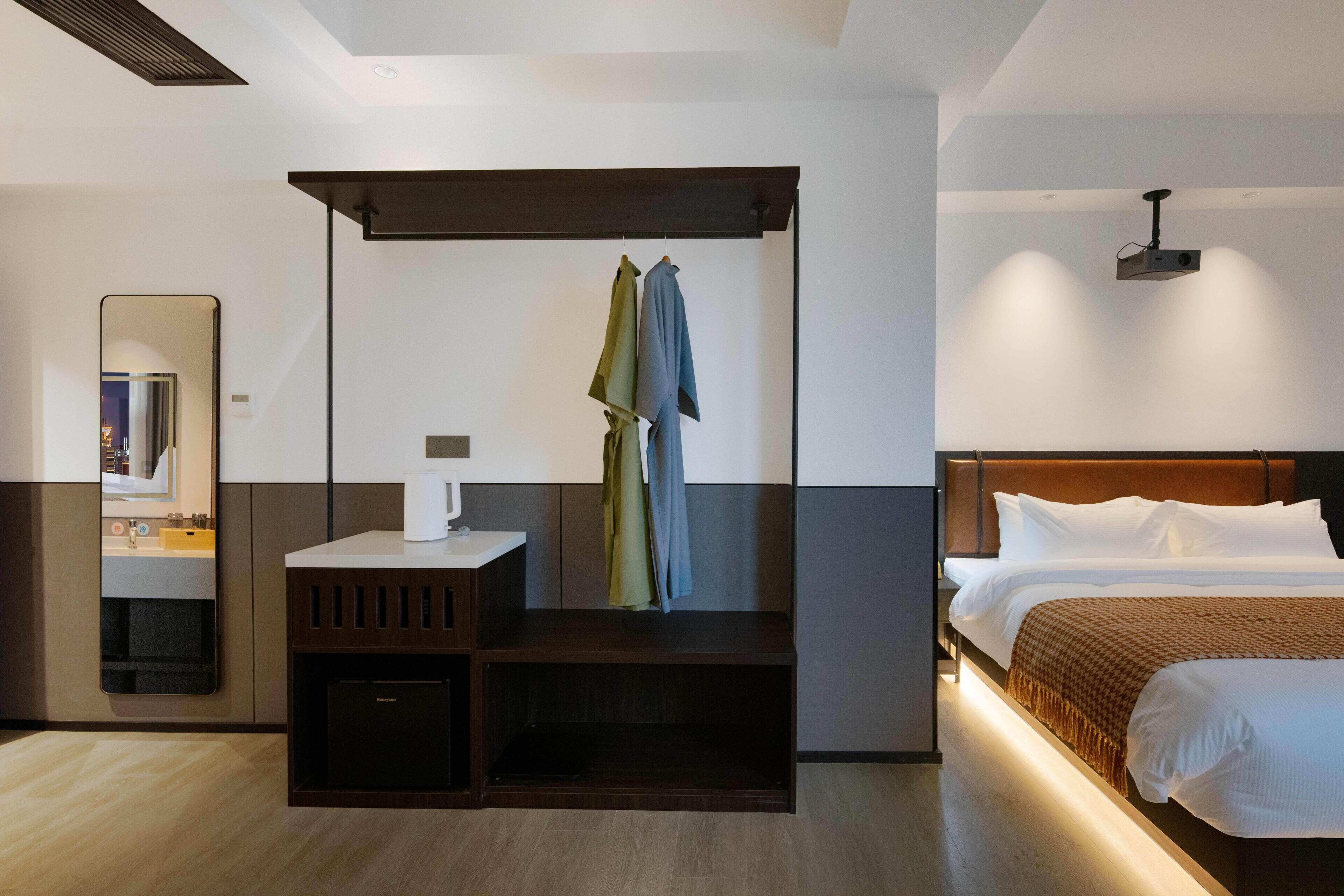 A modern hotel room with a bed, a bedside table with a lamp, a mirror, and a coat rack with clothes.