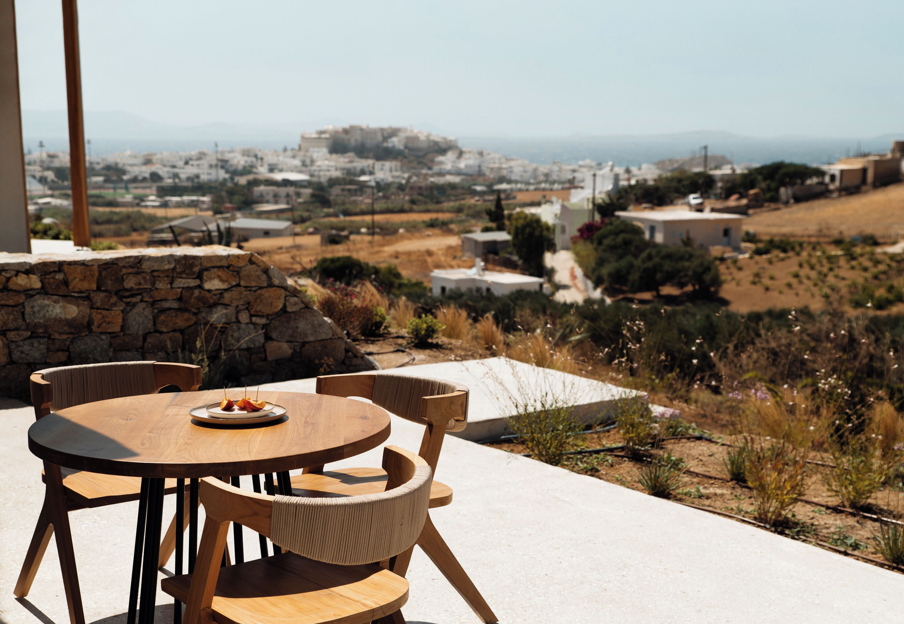 A round wooden table with chairs, a stone wall, and a view of a town with buildings and hills in the distance.