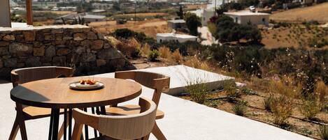 A round wooden table with chairs, a stone wall, and a view of a town with buildings and hills in the distance.