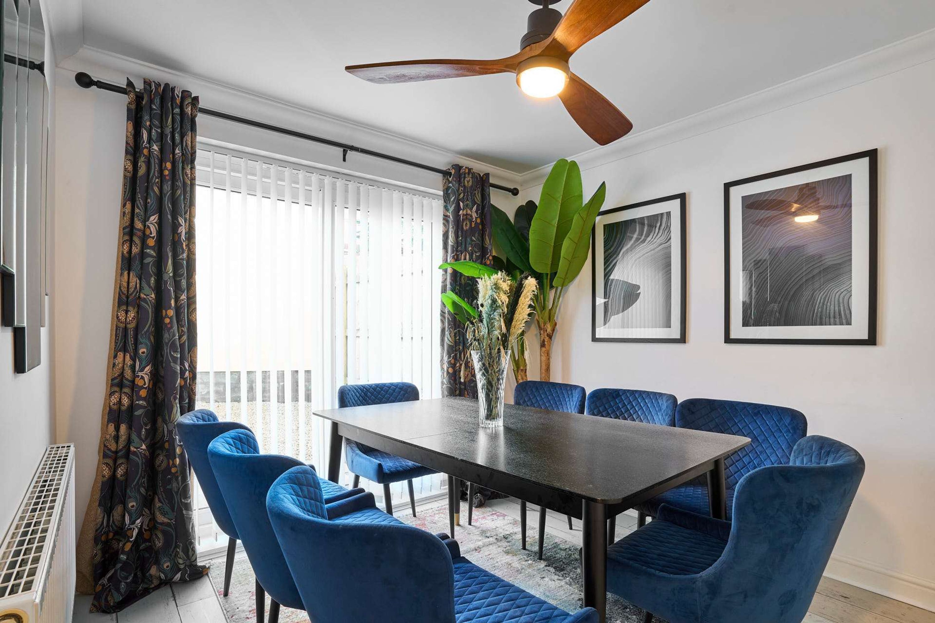 A dining area with a dark wooden table, blue upholstered chairs, a ceiling fan, and framed artwork on the wall.