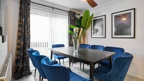 Un comedor con una mesa de madera oscura, sillas tapizadas en azul, un ventilador de techo y obras de arte enmarcadas en la pared.