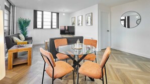Ein modernes Wohnzimmer mit einem Esstisch mit Glasplatte, orangefarbenen Sesseln, einem grauen Sofa, einem Fernseher und einem großen Fenster.