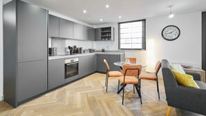 Eine moderne Küche mit grauen Schränken, eine Essecke mit einem runden Tisch und orangefarbenen Stühlen sowie ein graues Sofa mit einem gelben Kissen.