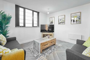 A modern living room with a grey sofa, a wooden coffee table, and a TV on a wooden stand. There are three framed artworks on the wall and a large window with curtains. - Host Stay Lockside House (Birmingham)