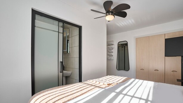 Un dormitorio moderno con cama, ventilador de techo, armario y baño visible a través de una puerta abierta.