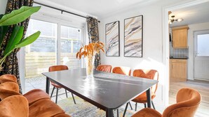 Un comedor con una mesa oscura, sillas anaranjadas y un jarrón con flores naranjas. Se observan ventanas grandes con cortinas, un ventilador de techo y obras de arte enmarcadas en la pared.