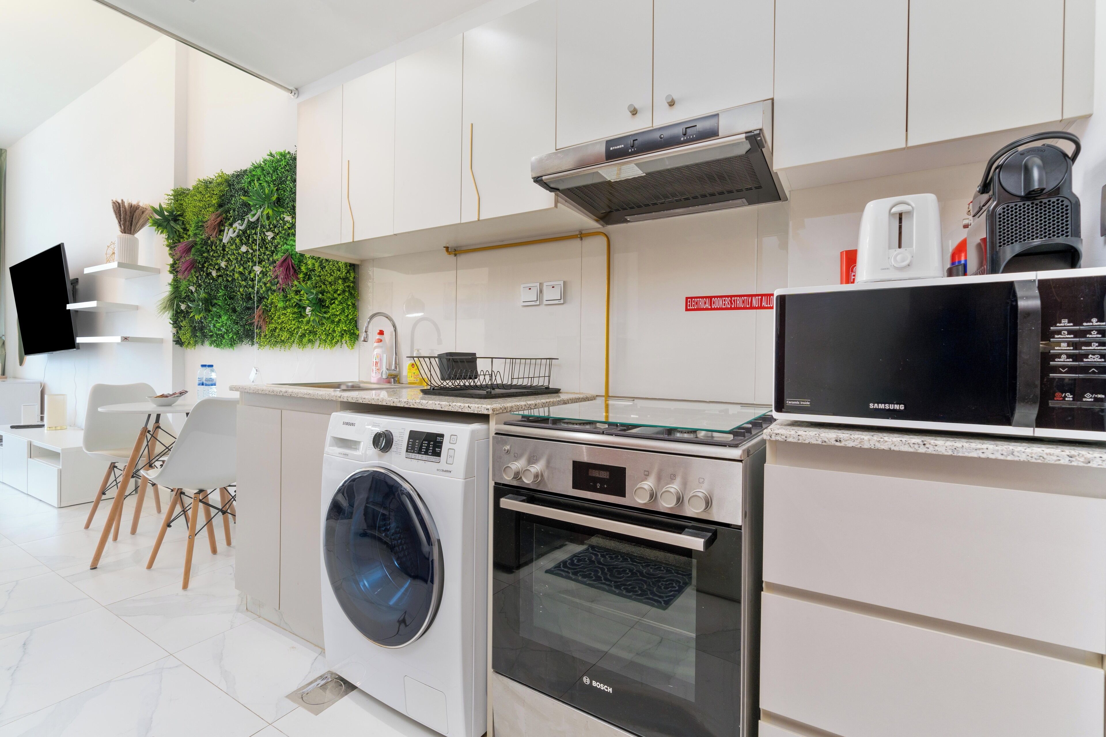 A modern kitchen with a washing machine, oven, microwave, and a green wall with plants.
