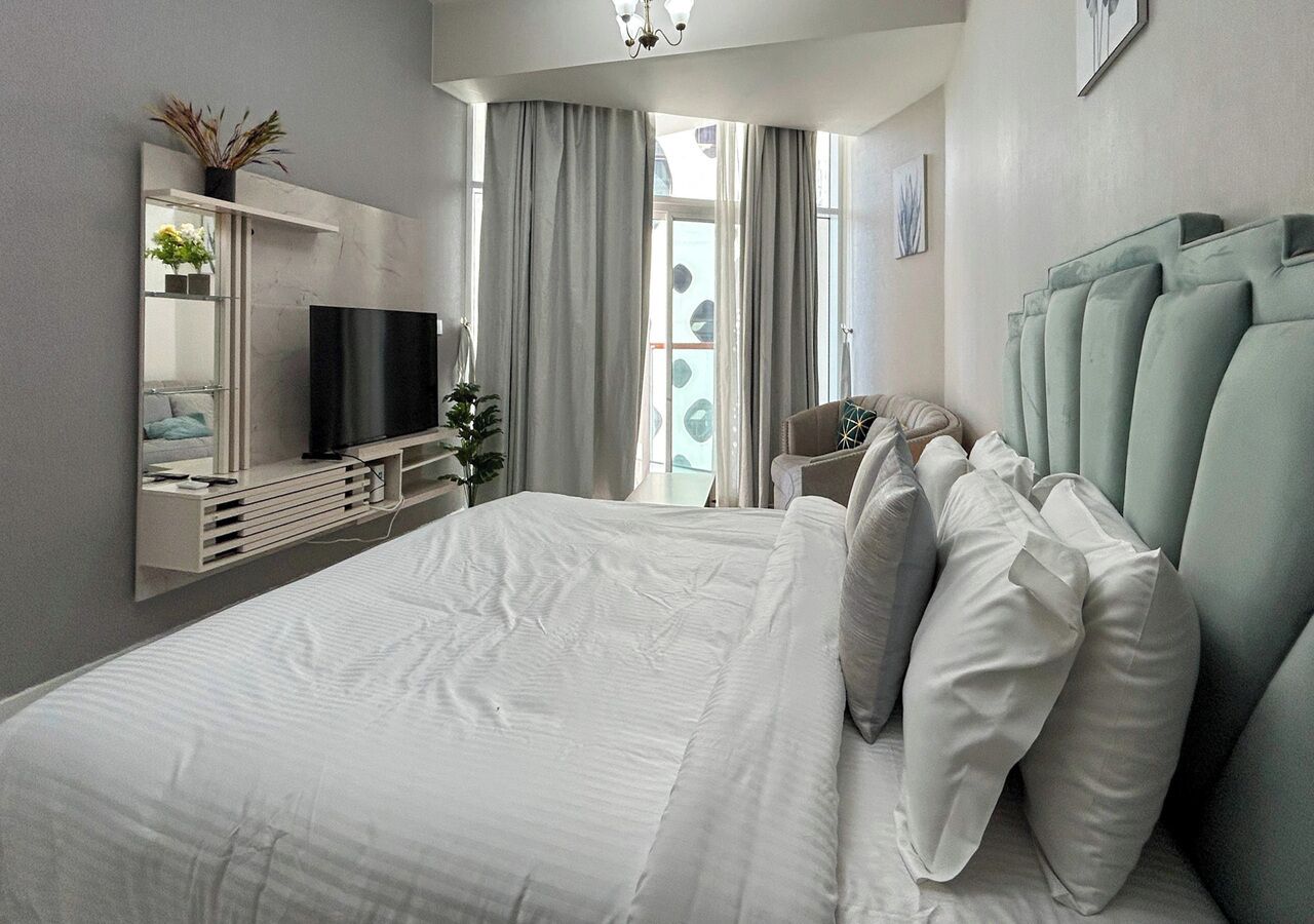 A modern hotel room with a large bed, a flat-screen TV, a bedside table, and a view of the outdoors through large windows.