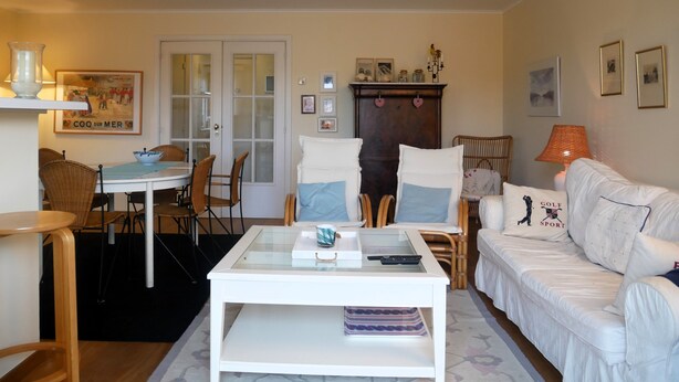 Un salon avec un canapé blanc, une table basse en verre, des fauteuils en osier, une table à manger avec des chaises et une cheminée.