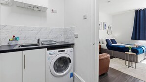A compact hotel room with a washing machine, a kitchenette, a bed, and a seating area. - Tigris - Cosy Studio Flat (Warrington)