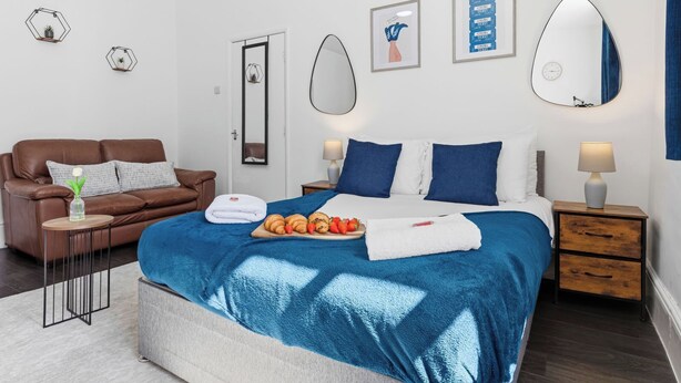 A bedroom with a bed, a brown leather sofa, a small table with a vase, and a nightstand with a lamp. - Tigris - Cosy Studio Flat (Warrington)
