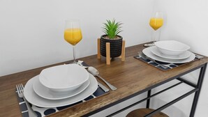 A dining table with white plates, cutlery, and glasses of orange juice, set against a white wall with a small potted plant. - Tigris - Cosy Studio Flat (Warrington)
