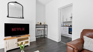 A modern living room with a TV, a leather sofa, a dining area with stools, and a kitchen in the background. - Tigris - Cosy Studio Flat (Warrington)