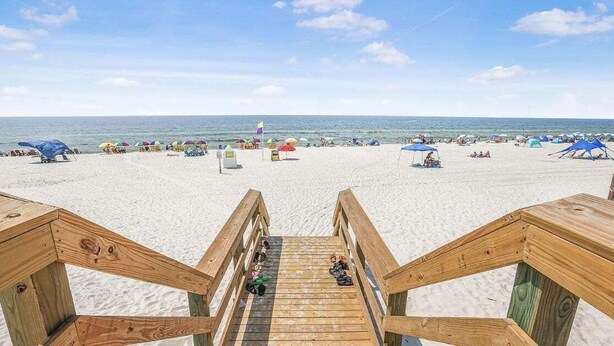 Eine Holztreppe führt zu einem Sandstrand mit Sonnenschirmen und Strandbesuchern.