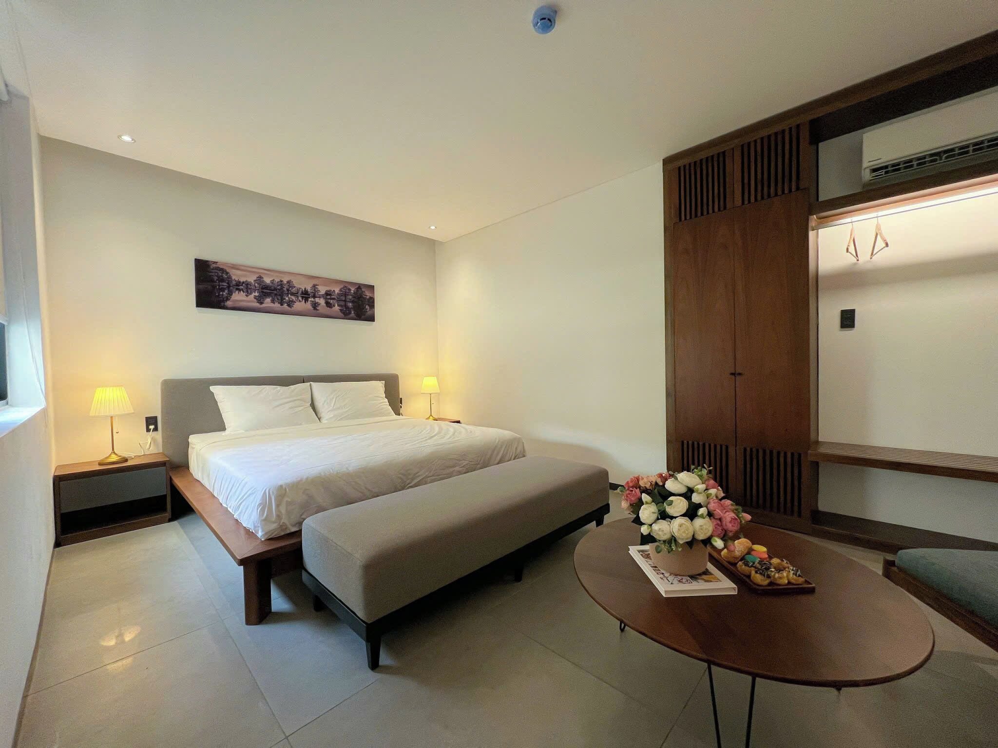 A hotel room with a bed, bedside tables, a bench, a round coffee table with flowers, and a wall-mounted air conditioning unit.