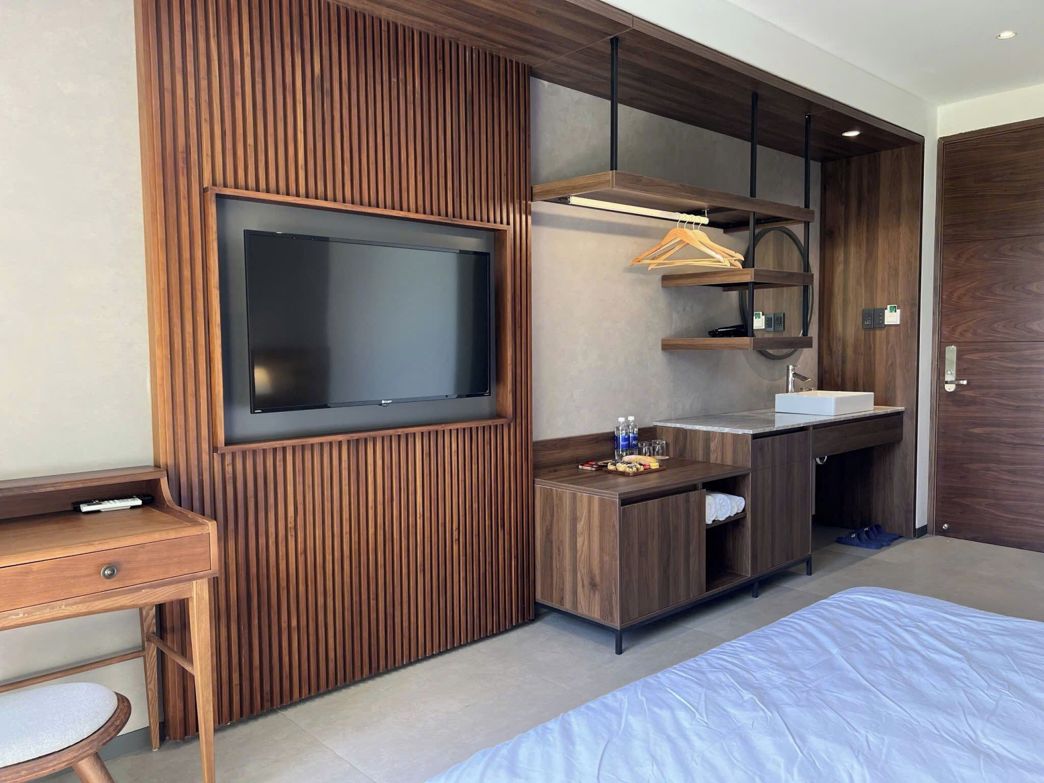 A modern bedroom with a TV, wooden paneling, a desk, a bed, a bathroom with a sink, and a mirror.