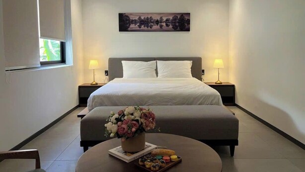 A modern hotel room with a large bed, two bedside lamps, a painting on the wall, a small table with a vase of flowers and pastries, and a window with blinds.