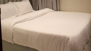 Un lit bien fait, avec une tête de lit grise et du linge de lit blanc, dans une pièce aux murs de couleur beige et ornée d’un rideau gris.