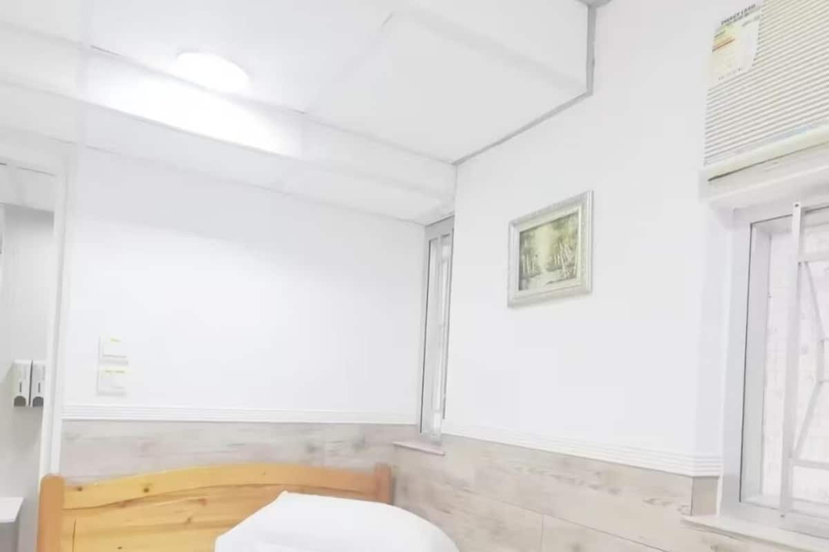 A neatly made bed with white linens in a room with a wooden headboard, white walls, and a window with blinds.