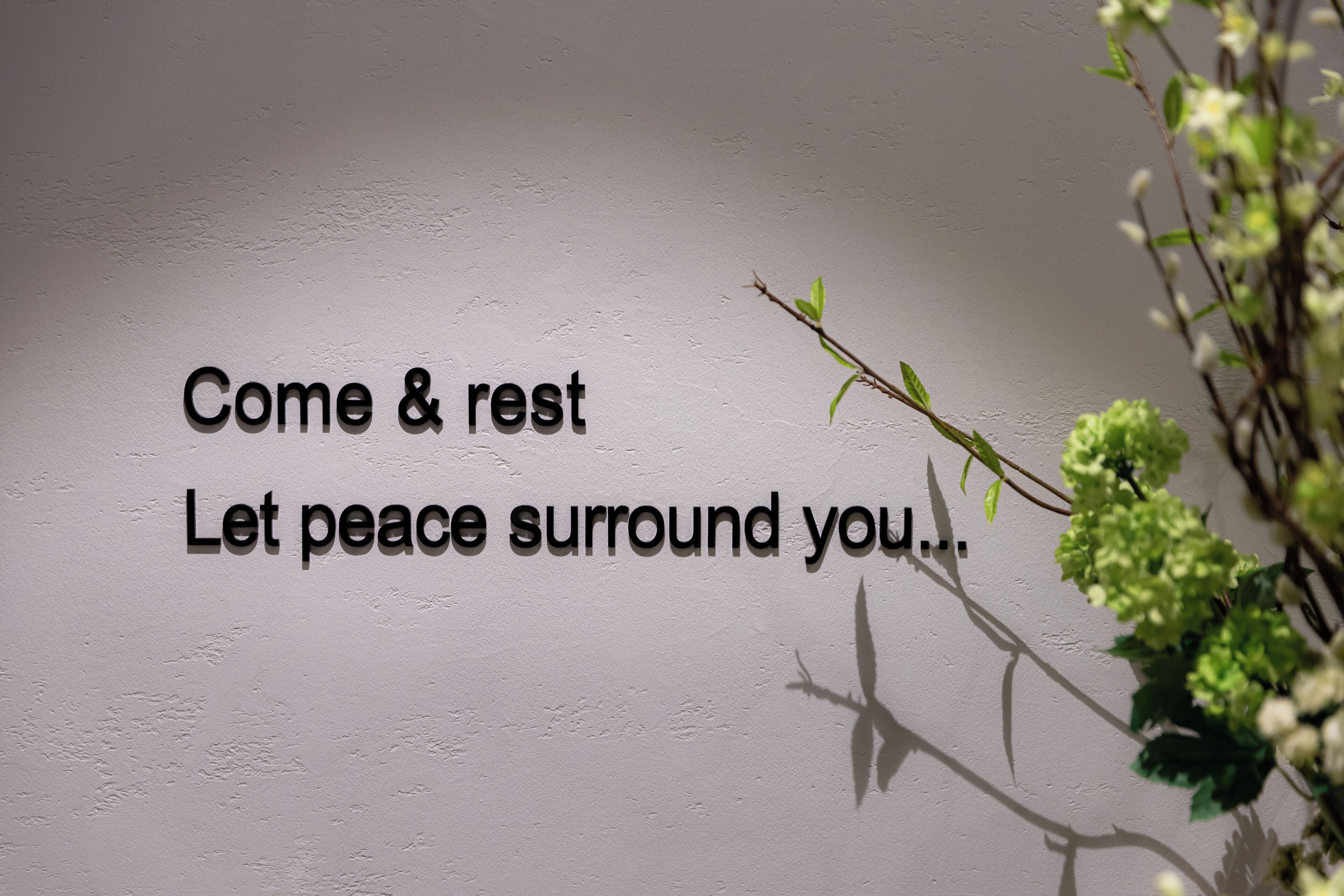 Una pared con el texto “Ven y descansa. Que la paz te rodee...” y una rama con hojas.