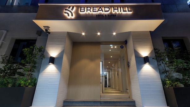 Das Außengelände des Bread Hill Hotel & Suites präsentiert sich mit einem modernen Design, einschließlich eines markanten Eingangs, Stufen, die zur Tür führen, und beleuchteter Beschilderung.