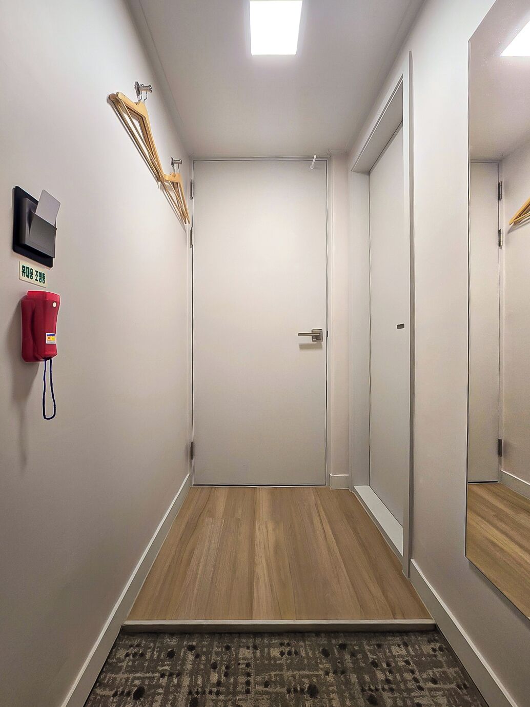 A hallway with a wooden floor, a fire alarm, a coat hook, and a door.