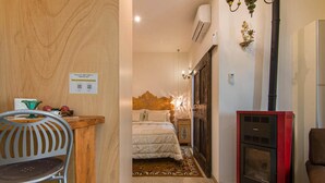 Una habitación de hotel pequeña con cama, un escritorio bajo con silla, un estufa de leña roja y un adorno de pared.