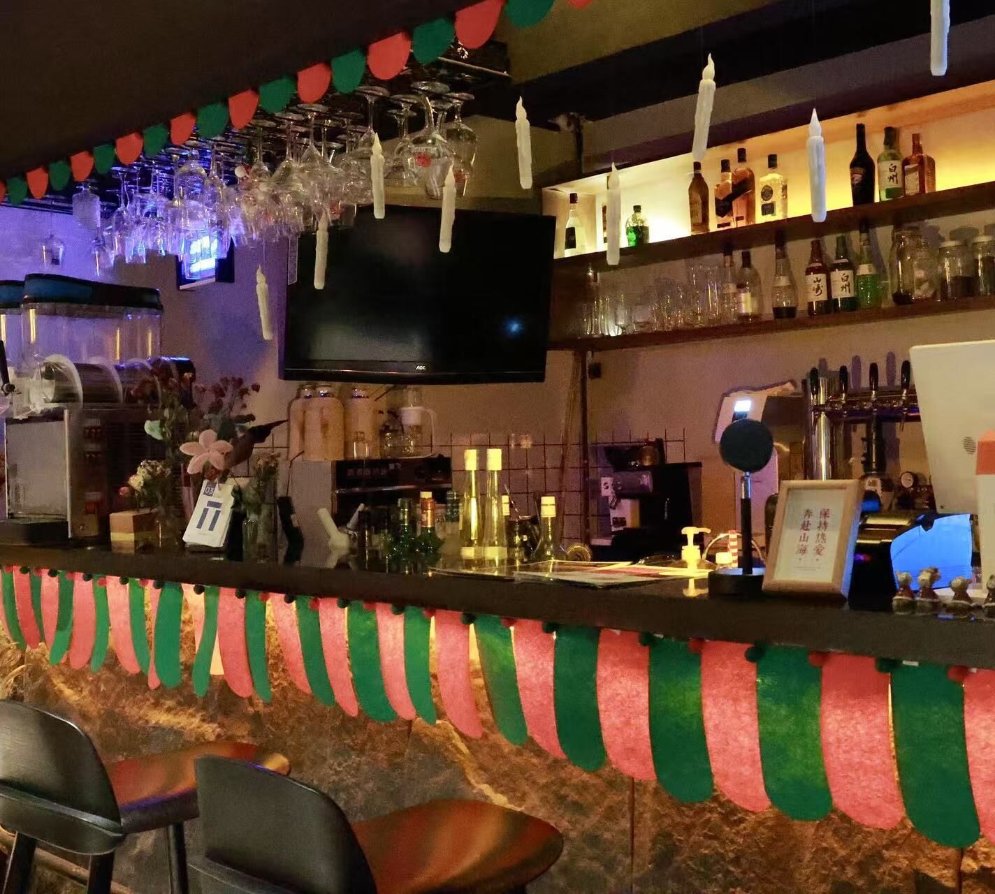 A bar area with a television, shelves with bottles, and hanging glasses.