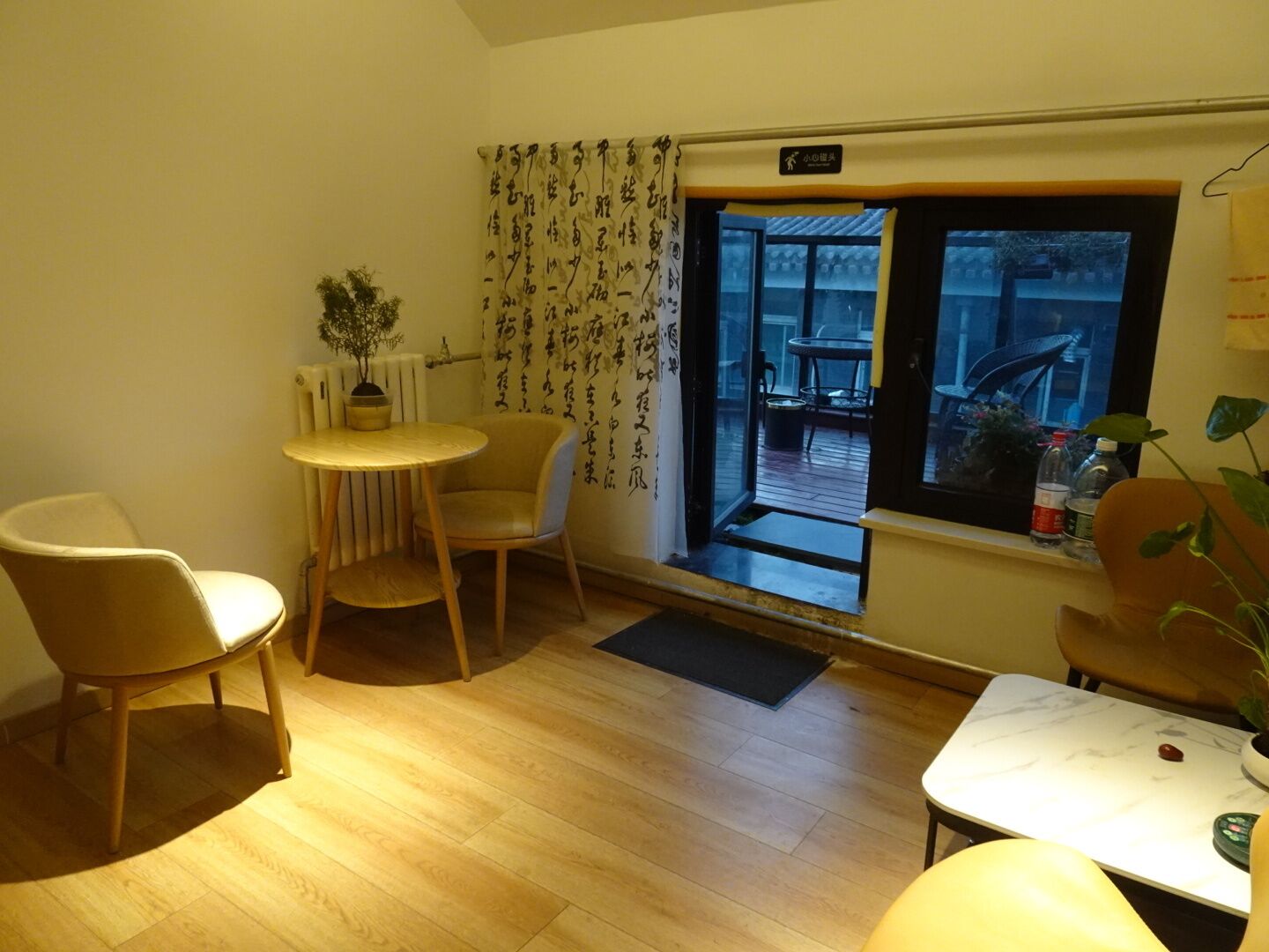 A room with a wooden floor, a small round table with chairs, a potted plant, and a window with a view of an outdoor patio.