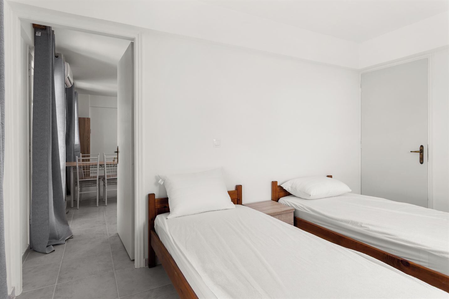 Un dormitorio con dos camas individuales, cabecera de madera, mesita de noche y cortina gris.
