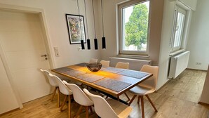 Een eetruimte met een houten tafel, witte stoelen, een gevlochten mand en een lijstwerk met kunst aan de muur.