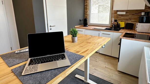 Een moderne keuken met een laptop op een houten tafel, witte kasten, een raam en een magnetron.