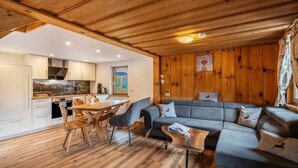 Een modern interieur met een houten plafond, een eethoek met een houten tafel en stoelen, een grijze bank en een keuken met witte kasten.