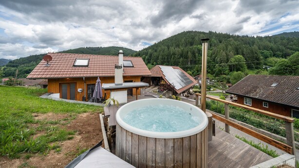 Een houten terras met een jacuzzi, een huis met een rood dak, en een beboste berg op de achtergrond.