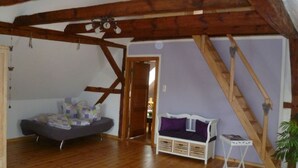 Een kamer met een schuine balkenplafond, een bed met een paarse deken, een witte bank en een kleine tafel.