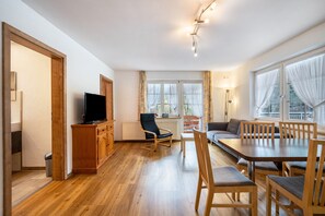 Un salon moderne avec un plancher en bois, un téléviseur, un canapé, une table à manger avec des chaises et de grandes fenêtres avec des rideaux.